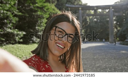 Cheerful, beautiful hispanic woman wearing glasses having fun making a confident selfie picture, smiling expression at meiji temple, tokyo's cultural shrine, capturing her happy moment.