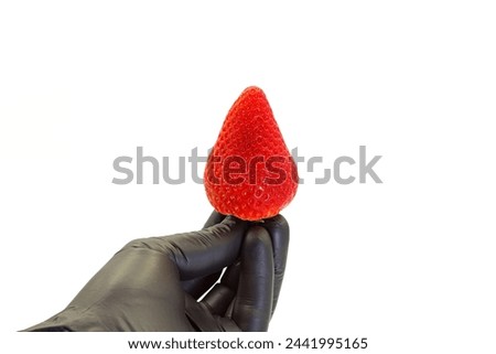 A black glove holding a strawberry on a white background. Royalty-Free Stock Photo #2441995165