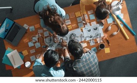 Top down aerial view of skilled designer hold logo while discuss with team. Aerial business group working together sharing idea about logo graphic design by using sticky notes and tablet. Symposium.