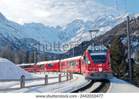 A Bernina Express travels thru Montebello Curve on a sunny winter day, with Morteratsch Glacier lying below Piz Bernina and snowy mountains in background in Pontresina, Graubünden, Switzerland, Europe Royalty-Free Stock Photo #2442010477