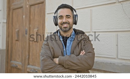 A smiling hispanic man with a beard wearing headphones outdoors in an urban city setting, exuding casual confidence.