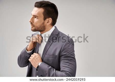 thoughtful businessman in suit adjusting his tie on grey background, corporate fashion concept Royalty-Free Stock Photo #2442019293