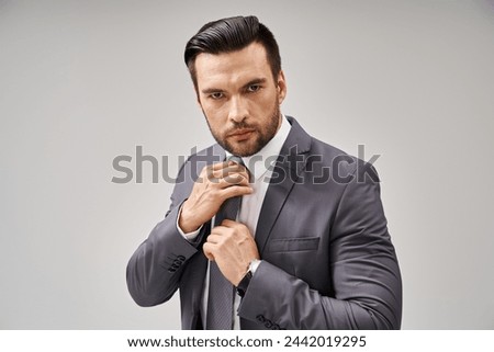 thoughtful man in formal wear adjusting his tie on grey background, corporate fashion Royalty-Free Stock Photo #2442019295