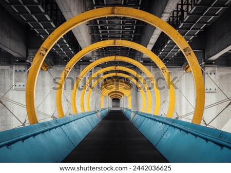The portal. Pedestrian footbridge under highway bridge. Symmetric diminishing perspective view of walkway with blue barriers and yellow metal structure surrounding it. Royalty-Free Stock Photo #2442036625