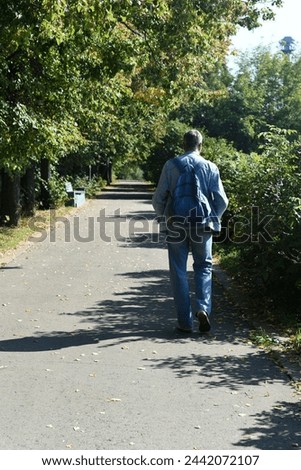 A man in a denim suit with a backpack on his back walks briskly along the alley of the park. Royalty-Free Stock Photo #2442072107