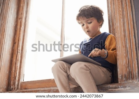 Home, tablet and serious kid on app, internet and typing for learning game by windowsill. Technology, child and boy in house for education, scroll and watch cartoon online on digital electronics