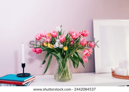 Modern interior design with tulip flowers bouquet in vase, white picture frame mockup, candles on white console on pink wall background. Poster design mockup. Minimalist Home decor. Selective focus