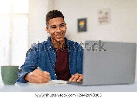 Happy African American teenager guy in blue shirt studying with grey laptop computer, taking notes, enjoying learning at home environment, free space Royalty-Free Stock Photo #2442098383