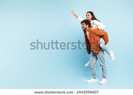 Joyful indian man giving piggyback ride to his young wife against blue studio backdrop, looking aside at free place for promotion text. Happy middle eastern couple having fun together. Copy space Royalty-Free Stock Photo #2442098407
