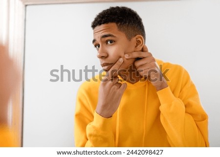 Black teenage guy in bright yellow hoodie checking his facial skin carefully in the mirror, focused on skincare and personal grooming, home interior Royalty-Free Stock Photo #2442098427