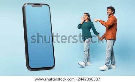 Wow mobile app. Excited middle eastern couple pointing at giant smartphone with mockup empty screen, recommending mobile application or website, standing near device on blue background. Panorama