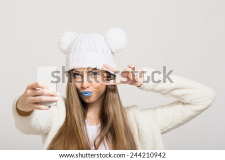 Closeup shot of cute teenage girl with blue lips and white knit hat taking a selfie with smart phone pouting and showing two fingers. Modern hipster young woman photographing herself with cellphone.