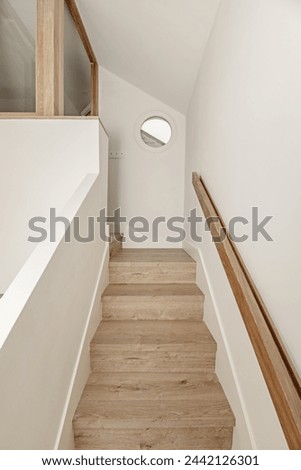 Stairs with wooden steps and oak handrails in the interior of a minimalist-style detached house with a porthole window in the attic Royalty-Free Stock Photo #2442126301