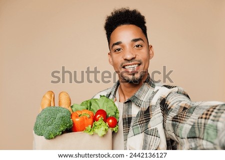 Young man wears grey shirt hold paper bag for takeaway mock up with food products doing selfie shot on mobile cell phone isolated on plain beige background. Delivery service from shop or restaurant