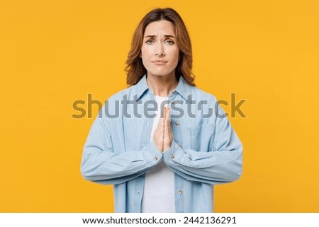 Young sad woman she wears blue shirt white t-shirt casual clothes hold hands folded in prayer gesture, begging about something isolated on plain yellow background studio portrait. Lifestyle concept Royalty-Free Stock Photo #2442136291