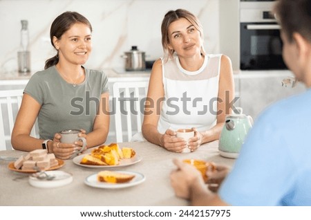 Smiling hospitable woman and young girl receiving male guest over cup of tea with sweets at home, sitting at cozy kitchen table and talking friendly.. Royalty-Free Stock Photo #2442157975