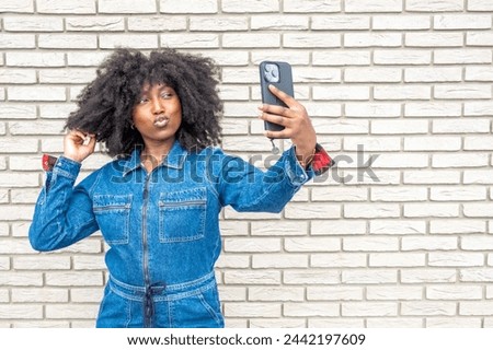 This vibrant image captures a young African American woman in a trendy denim outfit taking a selfie. Her playful pout and voluminous afro hairstyle stand out against the textured white brick wall