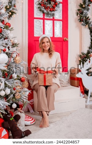 A young woman in a Santa hat with a gift in her hands sits on the porch next to the Christmas tree.