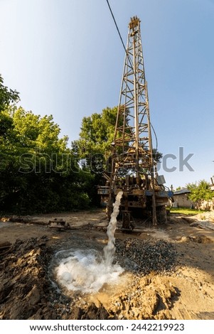 Old water pump truck extracts water from a hole in the ground at sunny summer day, surrounded by a natural landscape with trees, plants, and soil. Ultra-wide angle. Royalty-Free Stock Photo #2442219923