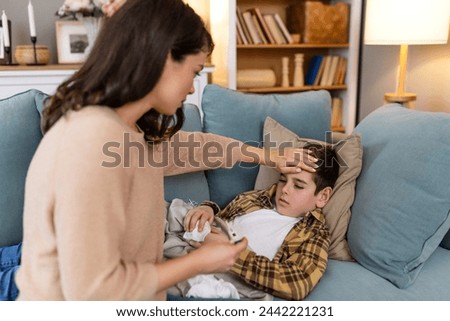 Young woman checking temperature with hand of her ill son. Mother checking temperature of her sick boy. Sick child lying on bed under blanket with woman checking fever on forehead by hand.