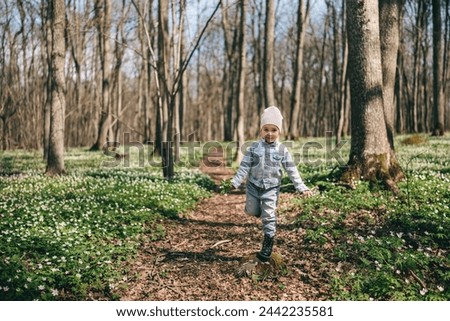 Portrait of adorable 3 years old girl in jeans jacket standing on the stump in the forest covered with anemones holding bouquet of forest flowers. Happy childhood 