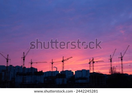 Tower cranes on building of  many-storeyed house stock photo. Six tall construction cranes stand out as dark silhouettes against the colorful sky.