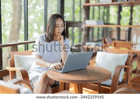 An Asian female student sits intently on her laptop working on a graduation project by the window on her desk. Wear casual clothes and explore the university's faculty library.