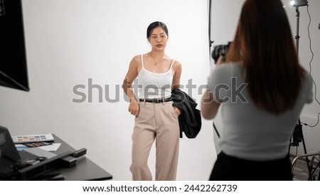 A professional female photographer is taking pictures of an attractive young Asian female model in casual wear, having a fashion photoshoot in a studio with professional lighting equipment.