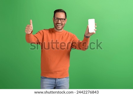 Cheerful young salesman showing thumbs up sign and smart phone's screen on isolated green background