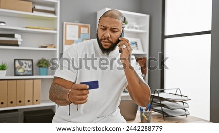 A concerned adult black man holds a credit card and talks on the phone in a modern office setting.