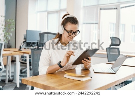 A tech-savvy woman in headphones enjoys interacting with a tablet, with a laptop nearby, in a sunlit, contemporary office space.

 Royalty-Free Stock Photo #2442292241