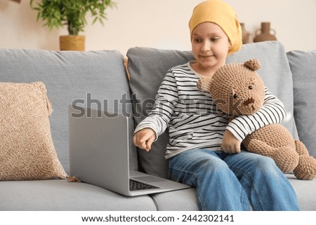 Little girl after chemotherapy with toy bear watching cartoons on laptop at home. International Childhood Cancer Day Royalty-Free Stock Photo #2442302141