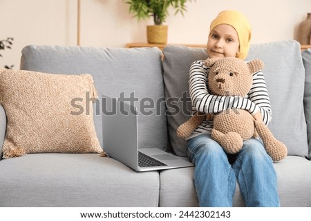 Little girl after chemotherapy with toy bear watching cartoons on laptop at home. International Childhood Cancer Day Royalty-Free Stock Photo #2442302143