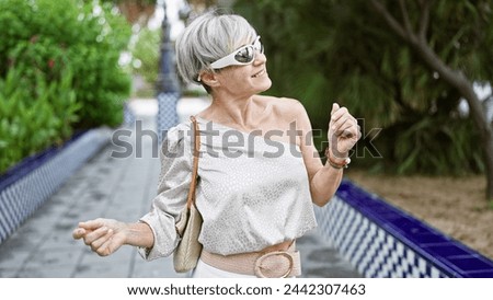 Elegant middle-aged woman with grey hair enjoying a sunny day in a green park, wearing sunglasses and a chic outfit. Royalty-Free Stock Photo #2442307463
