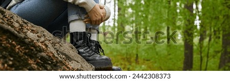 Cropped photo of female hiker hugging her legs wearing jeans and hiking boots in forest, banner