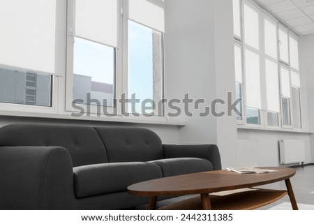 Comfortable sofa and table near large windows with white roller blinds indoors
