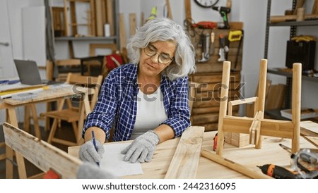 Mature woman in glasses plans woodworking project in a workshop Royalty-Free Stock Photo #2442316095