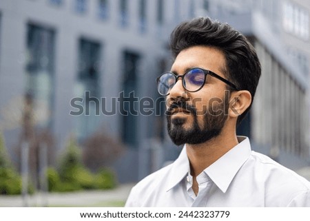 Close-up photo of a relaxed young Indian man wearing glasses and a white shirt sitting outside a business center ,with his eyes closed and resting.