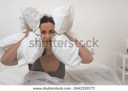 In anguish, she presses pillows to her ears, unable to sleep due to noisy neighbors, afflicted by a throbbing headache and insomnia.  Royalty-Free Stock Photo #2442350171