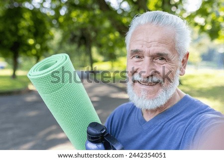 Close-up photo of an older smiling gray-haired man standing in a park, holding a mat and a bottle of water, smiling and talking to the phone camera.