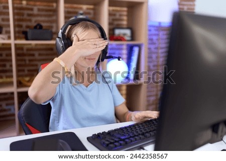 Young caucasian woman playing video games wearing headphones covering eyes with hand, looking serious and sad. sightless, hiding and rejection concept  Royalty-Free Stock Photo #2442358567