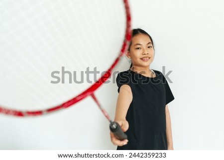 Portrait of an Asian child girl holding a badminton racket with the white wall background, the girl pointing the badminton racket to the camera.