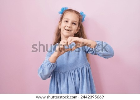 Young little girl standing over pink background smiling in love showing heart symbol and shape with hands. romantic concept. 