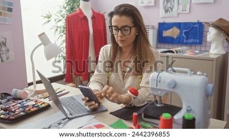 A focused woman tailor in an atelier checks her phone amid sewing equipment, fashion designs, and mannequins.