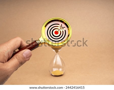 Short-term business goals encompass work that helps an organization reach its mid-term goals concept. Target icon in gold magnifying glass lens in hand while zoom in at hourglass on brown background. Royalty-Free Stock Photo #2442416415