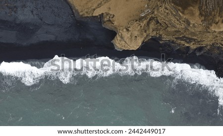 A bird's-eye view captures the dramatic interaction between the tumultuous North Atlantic waves and Iceland's famed black sand shores, bordered by rugged terrain. Royalty-Free Stock Photo #2442449017