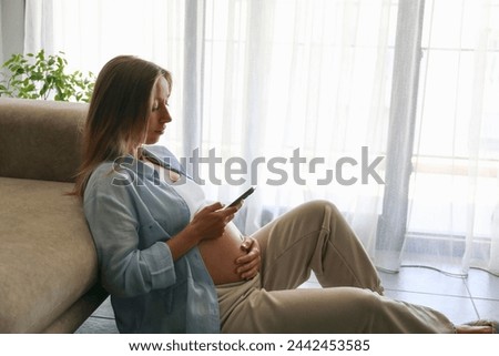 Pregnancy tracking app concept. Young pregnant woman sitting on the floor and checking her phone. Close up, copy space, background.