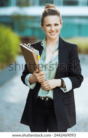 smiling modern middle aged business woman near business center in black jacket with documents and folder. Royalty-Free Stock Photo #2442469093
