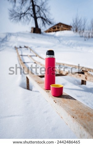 Pink thermos with cup standing on board on winter landscape background, frosty spring morning, hot coffee in thermos, camping utensils. High quality photo