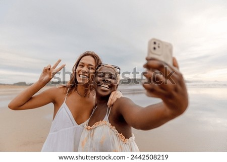 Two female friends of different races (Latina and black) using a smart phone to take pictures together on a beach during a summer trip.